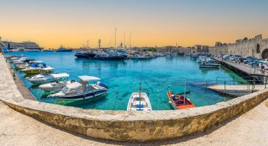 RHODES, GREECE - 30 Haziran 2022: İtalyan döneminden kalma yatlar, botlar ve tarihi ortaçağ duvarları, Mandraki limanındaki deniz gezinti alanı boyunca.