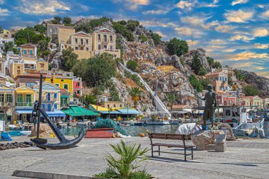 SYMI, GREECE-JULY 3 Temmuz 2022: Metal çapa heykeli ile kentin gezinti alanı, 20. yüzyılın başlarında Suriye 'de sünger dalgıçlarının başı olan Stathis G. Hatzis' in heykeli ve çok renkli evler.