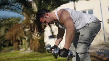 An athlete with a beautiful body and large muscles raises his hands with dumbbells in nature. High quality 4k footage
