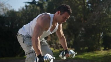 An athlete with a beautiful body and large muscles raises his hands with dumbbells in nature. High quality 4k footage