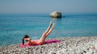 Bir kadın enerjik bir şekilde arka planda açık mavi su olan güzel bir plajda çeşitli egzersizler yapıyor.