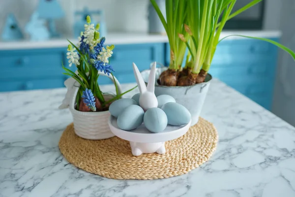 Blue kitchen interior with furniture. Stylish cuisine with flowers in vase. Wooden kitchen in spring decor. Cozy home decor. Kitchen utensils, dishes and plate on table. kitchen island in dining room, bunny figurine. Easter concept