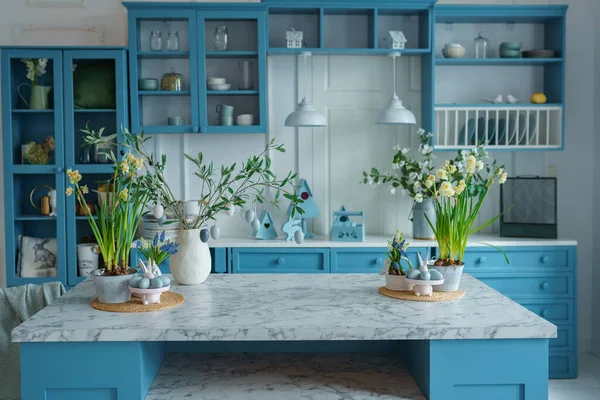 Blue kitchen interior with furniture. Stylish cuisine with flowers in vase. Wooden kitchen in spring decor. Cozy home decor. Kitchen utensils, dishes and plate on table. kitchen island in dining room, bunny figurine. Easter concept