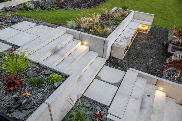 Residential House Backyard Garden with Concrete Stairs and Grill Area Surrounded by Lawn and Flower Beds Decorated with Black Pebbles.