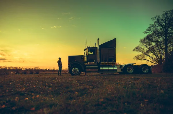 Semi Truck Driver in Front of His Vehicle During Scenic Countryside Sunset. Transportation Industry Theme.