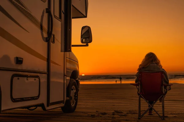 女人平静地坐在沙滩上的椅子上 看着她的丈夫在海里与狗玩耍 现代房车靠岸了 日落景致 — 图库照片