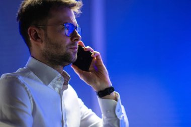 Caucasian Entrepreneur Having a Late Business Phone Call in His Office. Blue Background with Copy Space.