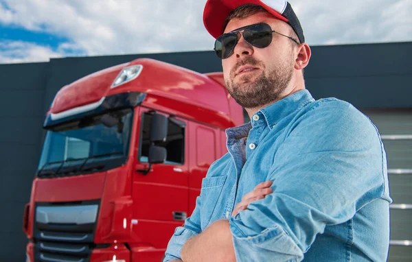 Closeup of Proud Caucasian Professional Trucker and His Red Semi Tractor Parked at Logistic Center Parking Lot in the Background.