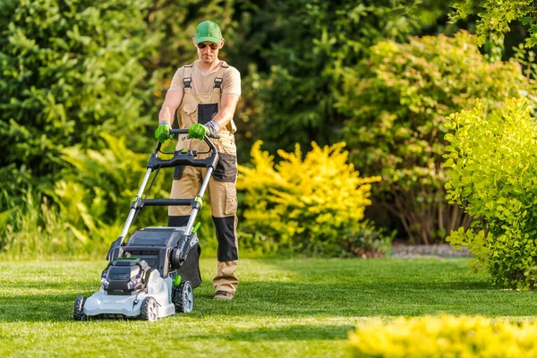 Professional Caucasian Gardener Mowing the Lawn with Battery-Powered Mower During Residential Garden Summer Maintenance.
