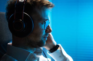 Caucasian Man in His 30s Listening Podcast on His Computer Using Professional Wireless Headphones. Dark Blue Illumination Scene. 