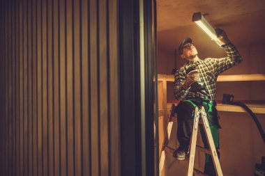 Caucasian Electrician in His 40s Installing LED Light Fixture in the Shed Ceiling. Small Wooden Building Illumination clipart