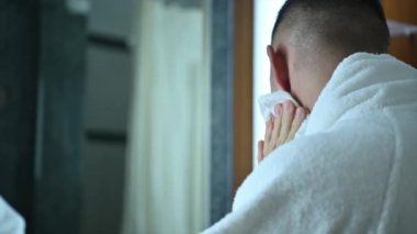 40 'lı yaşlarda beyaz bornoz giyen, yüzünü yıkayan ve sabah rutininde havluyla silen beyaz bir adam..