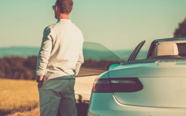 Günbatımının tadını çıkaran, modern beyaz üstü açık arabasının yanında duran zengin beyaz adam. Mutlu Yaşam.