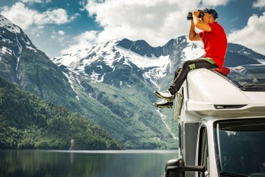 Caucasian Man in His 40s Exploring Scenic Norwegian Nature Using Binoculars While Seating on a Roof of His Camper Van RV Recreational Vehicle. Norway, Europe. clipart