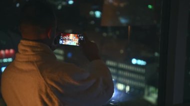 Beyaz bir adam otel odasının penceresinden Akşam Şehir Manzarası 'nın fotoğrafını çekiyor. İş Gezisi Teması.