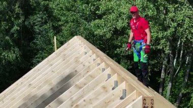 Çatı işçisi ahşap bir çatının tepesinde dikiliyor, dikkatlice dengeliyor. Çevreye bakıyor ve bölgeyi inceliyor gibi görünüyor..