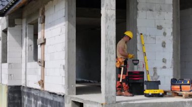 Sert şapka ve yansıtıcı yelek giymiş bir inşaat işçisi inşaat alanında yeni dökülmüş beton bir duvarın yanında duruyor. İşçi duvarı yakından inceliyor, muhtemelen yapılan işin kalitesini inceliyor..