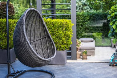 Bir siyah hasır yumurta sandalyesi, yeşil alanlarla çevrili bir verandadaki metal tezgahta asılı duruyor. Sandalye arka planda gri bir veranda ile büyük bir saksı çalısı önünde asılı.