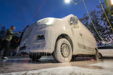 Bir işçi akşam araba yıkamada bir minibüse kalın köpük sürer. Kısmen köpükle kaplı araç, tepedeki ışıklarla aydınlatılıyor. Gündüz geceye geçerken canlı bir atmosfer yaratıyor..