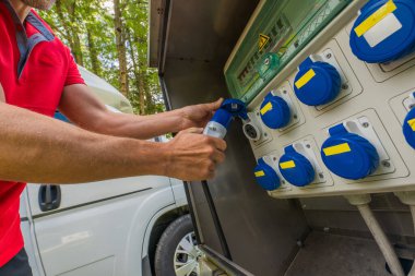 Bir kişi, ağaçlarla çevrili bir kamp alanındaki elektrik destek kablosunu bir elektrik kutusuna bağlıyor. Sahne, doğada rahat bir konaklama sağlamak için elektrik erişimi hazırlıklarını yakalıyor..