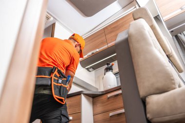 Turuncu gömlekli ve kepli bir işçi modern bir eğlence aracının mutfak alanını temizlemeye odaklanmış durumda. Mutfakta ahşap dolaplar ve aletler yer alıyor, temiz ve işlevsel bir yaşam alanı vurgulanıyor.