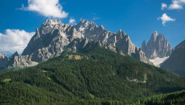 Seksi, İtalya Dolomitlerin yükselen zirveleri beyaz bulutlarla benekli canlı mavi gökyüzüne karşı görkemli bir şekilde yükseliyor. Dağın tabanındaki yemyeşil orman İtalyan Alplerinin doğal güzelliğini gözler önüne seriyor ve nefes kesici bir manzara yaratıyor.