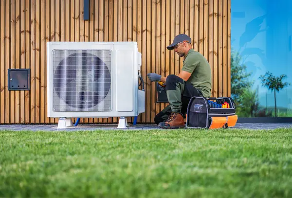 stock image A technician is servicing an air conditioning unit located beside a modern home. Dressed in work attire, he uses tools from an organized toolbox while seated on the grass. The setting features a wooden wall and bright blue windows, indicating a well-