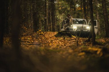 Kamuflaj kıyafeti giymiş bir yürüyüşçü öğleden sonra saatlerinde güzel sonbahar ormanlarına hayran olan beyaz bir SUV 'nin üzerinde oturur..