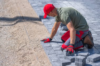 A worker is carefully laying down pavers on a prepared gravel base, focusing on alignment and precision. clipart