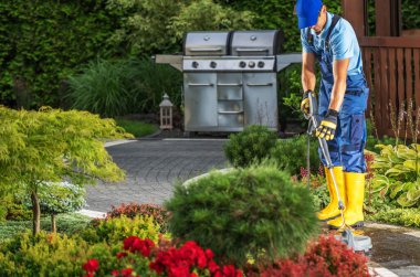 Sarı çizmeli bir bahçıvan güzel bir bahçedeki çalıları rengarenk çiçekler ve ızgarayla buduyor..