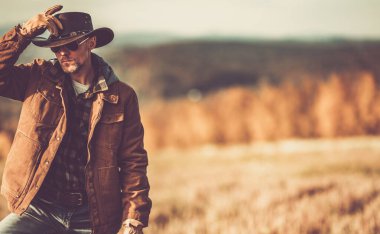 Bir adam, parlak sonbahar yapraklarıyla çevrili güneşli bir alanda dururken kovboy şapkasını düzeltir..
