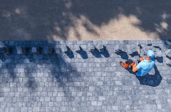 stock image A construction worker carefully positions pavers on a pathway, surrounded by tools and materials in bright sunlight.