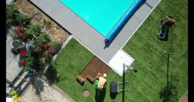 Bir insan çimenlik üzerine çalışarak ve arka bahçedeki bir yüzme havuzunun yanında malzeme düzenleyerek çalışır..