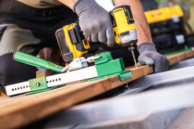 A carpenter is using a deckboard wrench and power drill to secure wooden boards on a construction site patio floor. clipart