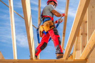 Bir inşaat işçisi dikkatli bir şekilde ahşap kirişlerde yürüyor, güvenlik teçhizatı ve aletleri giyiyor, güneşli bir inşaat alanındaki görevlerini tamamlarken.