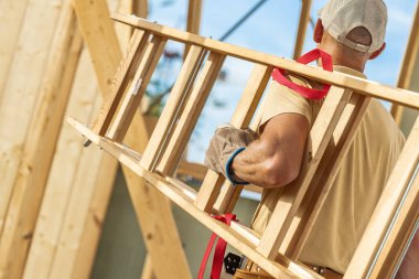Bir inşaat işçisi, kısmen inşa edilmiş bir binanın içinde çalışırken ahşap bir merdiven taşıyor. Tadilatın yoğun bir gününü gösteriyor..