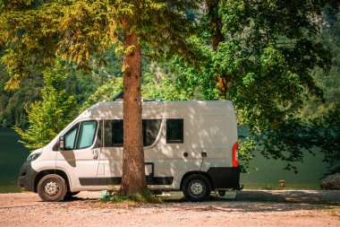 Konforlu bir karavan, sakin bir gölün yanına park edilmiş. Yüksek ağaçlar ve sıcak öğleden sonra ışığı altında canlı yapraklarla çerçevelenmiş. Huzurlu bir kaçış atmosferi yaratıyor..