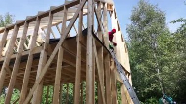 Bir kişi ahşap bir çerçeve inşa ediyor, merdivene tırmanıyor ve ağaçlar ve doğal manzarayla çevrili olarak üst kısımlarda çalışıyor..