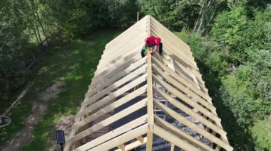 İki inşaatçı, berrak bir mavi gökyüzü altında, sakin bir ormanlık alanda ahşap bir çatı iskeleti kuruyor, marifetli işçilik ve takım çalışması sergiliyor..