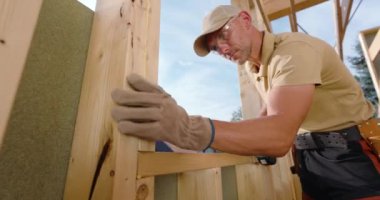 Profesyonel bir marangoz tahtaları titizlikle ahşap bir yapıya bağlar açık mavi gökyüzünün altında, işçilik ve projeye adanmışlık sergiler..