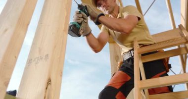 Bir inşaat işçisi, tahta kirişleri birleştirmek için ustalıkla bir matkap kullanırken açık gökyüzünün altındaki bir merdivende duruyor..