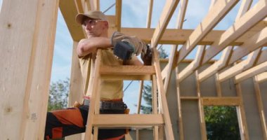 Bir inşaat işçisi, açık havada, güpegündüz, ahşap bir iskelet üzerinde çalışırken, bir merdivenin üzerinde ustalıkla elektrikli aletleri çalıştırır..