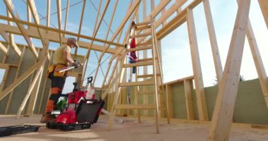 Bir inşaat işçisi, bir evin içindeki tahta kirişler üzerinde çalışırken bir merdiven üzerinde elektrikli bir alet kullanır. Parlak güneş ışığı yoğun atmosferi vurgular.