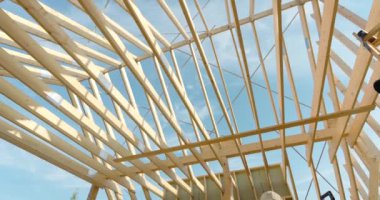 İşçiler, açık çatı kirişleri olan ahşap bir yapı inşa ediyorlar. Açık mavi gökyüzünün altında işçilik sergiliyorlar. Alan aktivite ve ilerlemeyle uğulduyor..