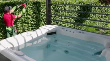 Bahçıvanın biri, özel bir bahçede jakuzinin yanındaki yemyeşil çitleri buduyor ve güneşli bir öğleden sonranın tadını çıkarıyor..