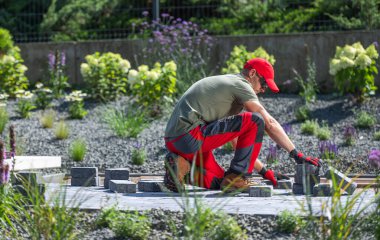 A gardener is meticulously arranging gray paving stones in a beautifully landscaped garden, surrounded by greenery and blooming flowers under bright sunlight. clipart