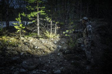 Bir birey gece karanlık bir ormanda dikkatle geziniyor, ağaçlar ve araziler arasındaki yolu aydınlatmak için bir el feneri kullanıyor, gizli detayları ortaya çıkarmak için..