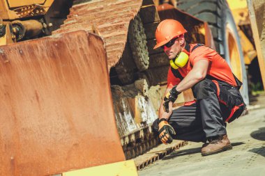 Turuncu gömlekli ve miğferli bir inşaat işçisi, şantiyede parlak bir günde çömelmiş ağır makineleri inceliyor..