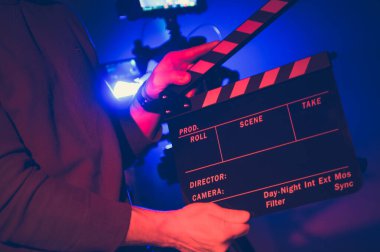 Bir film yapımcısının elinde prodüksiyon detaylarıyla etiketlenmiş bir ilan tahtası var. Canlı ışıklandırmada bir gece çekimine hazırlanıyor. Sahne arkası aktivitelerini sergiliyor..