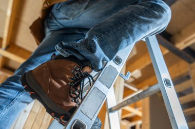 Bir işçi, sağlam botlar ve kot pantolon giyen, parlak tepelik ışıkları altında ev yenileme işleriyle uğraşan bir merdivene dikkatlice yerleştirilir..